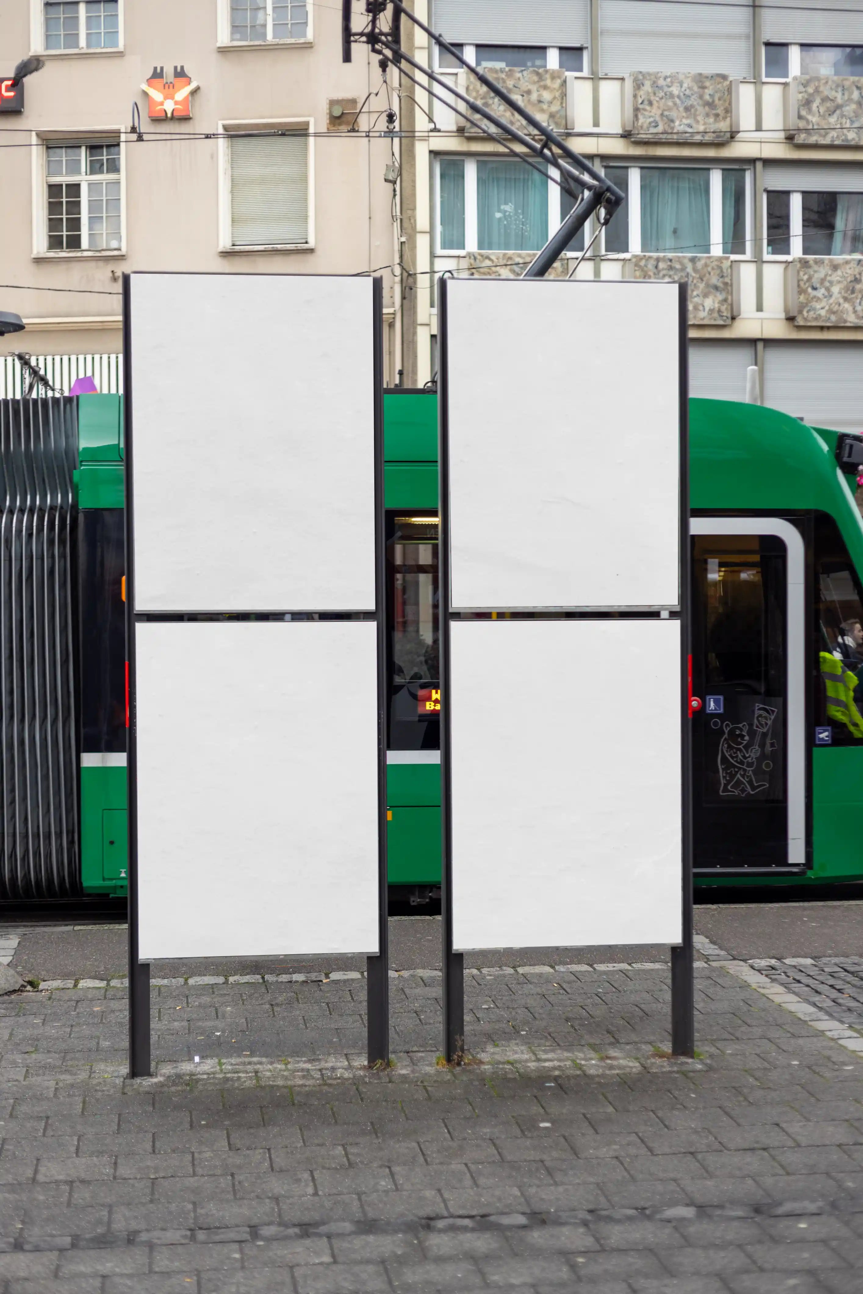 Four blank DIN poster mockups on two freestanding street advertising displays at Claraplatz in Basel, with a green tram, city architecture and urban pavement in the background. Ideal for clean poster series, campaign layouts and public transport advertising previews.