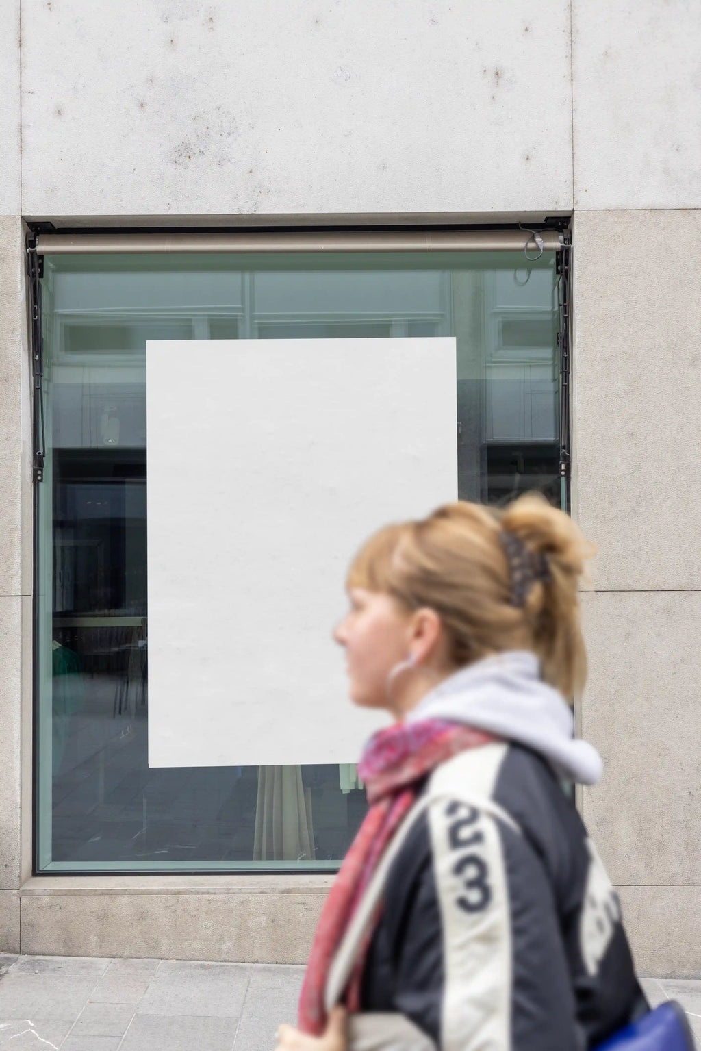Blank white poster mockup placed inside a shop window on Freie Strasse in Basel. A blurred person passes in front of the glass, with reflections, a light stone storefront and an urban shopping street setting for realistic retail poster presentations.