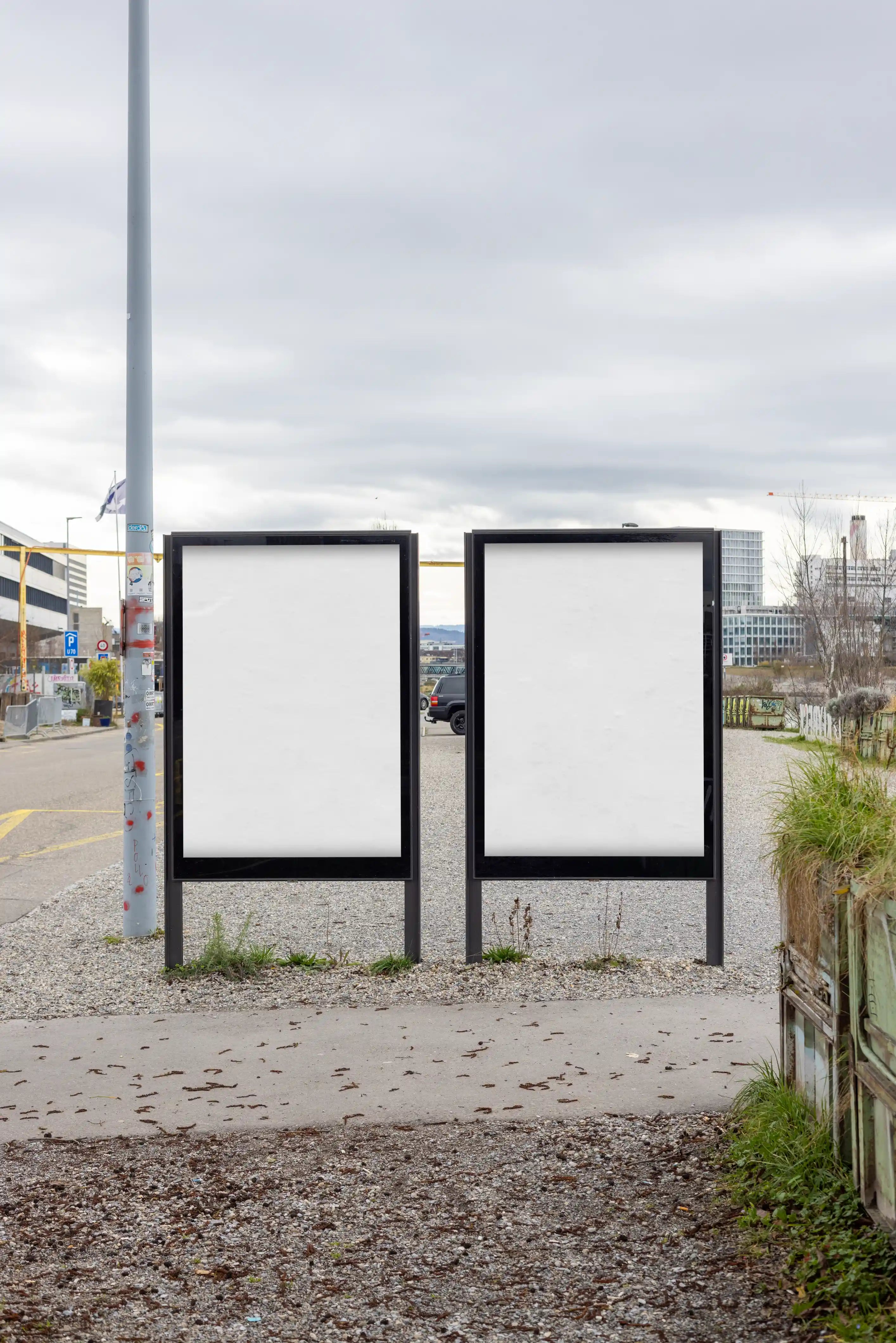 Two blank white city light poster mockups in DIN format standing near the waterfront at Gannet / Holzpark Klybeck in Basel. The scene features black display frames, gravel ground, grass, cloudy sky and urban surroundings, created for realistic outdoor poster advertising and campaign design previews.