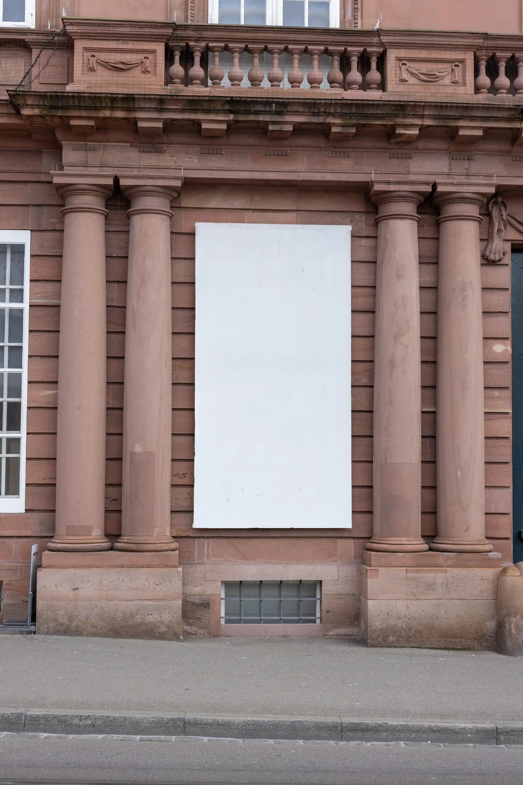 Blank vertical 1:2 banner mockup on the facade of the Historisches Museum Basel, Haus zum Kirschgarten. The white banner hangs between historic stone columns on a reddish sandstone building, with architectural details, sidewalk and street curb visible in the scene.