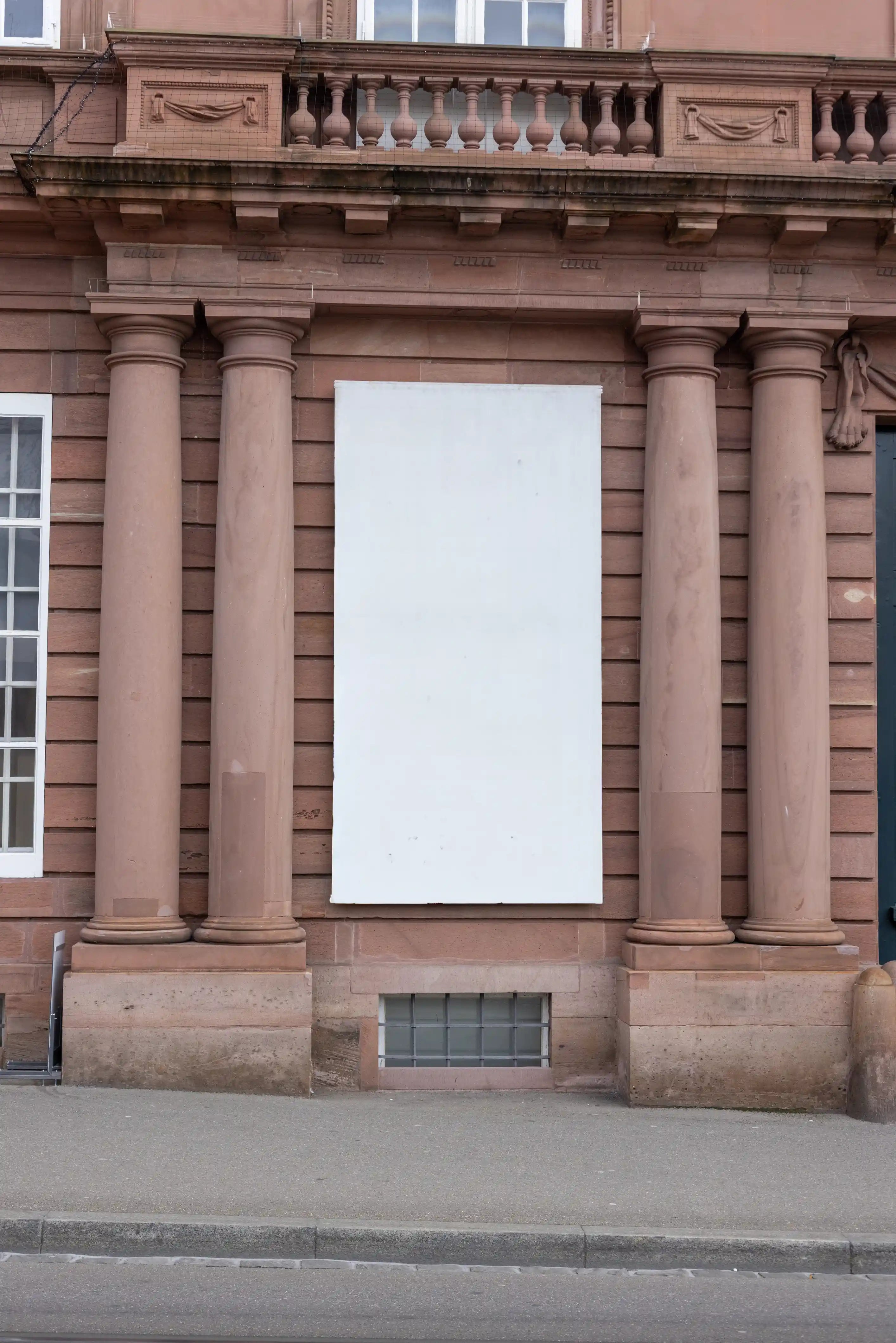 Blank vertical 1:2 banner mockup on the facade of the Historisches Museum Basel, Haus zum Kirschgarten. The white banner hangs between historic stone columns on a reddish sandstone building, with architectural details, sidewalk and street curb visible in the scene.