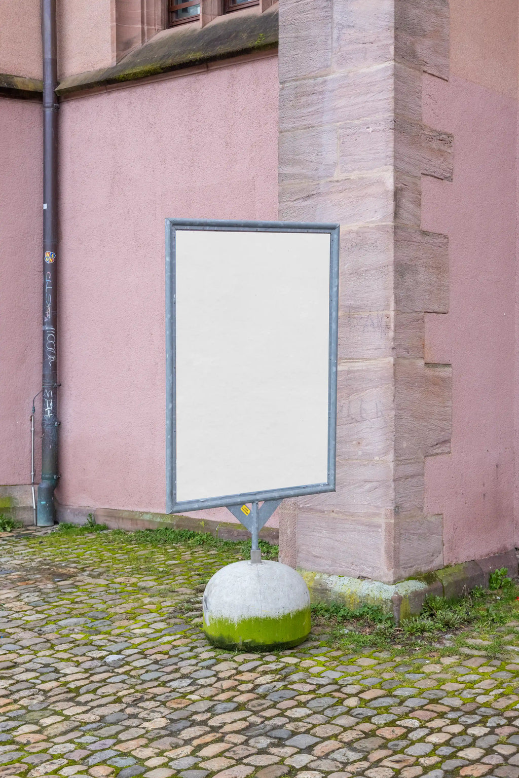 Blank vertical DIN poster mockup at Kaserne Basel near Ausstellungsraum Klingental, mounted on a freestanding metal sign display. The poster stands in front of a pink textured building wall with stone architecture, cobblestones, green moss and natural outdoor lighting.