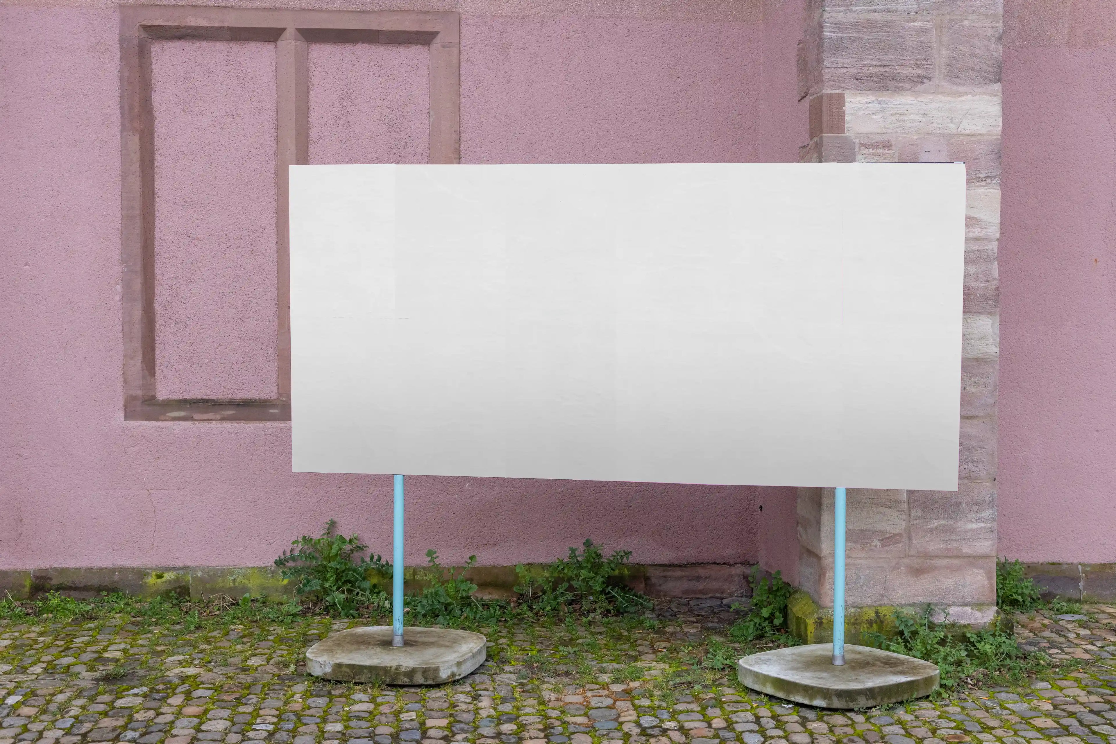 Blank wide poster mockup at Kaserne Basel near Ausstellungsraum Klingental, placed on a freestanding outdoor board in front of a pink wall and stone building corner. The scene includes cobblestone pavement, greenery and two blue support poles, ideal for large campaign previews and multi-poster layout presentations.
