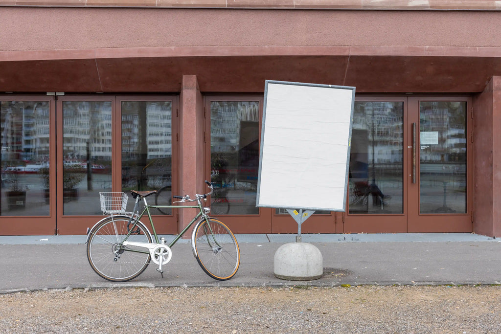Blank white DIN poster mockup on a freestanding metal display at Kaserne Basel near the Rhine. A bicycle is parked beside the sign, with reflective glass doors, reddish-brown building details, gravel ground and a quiet urban riverside atmosphere.