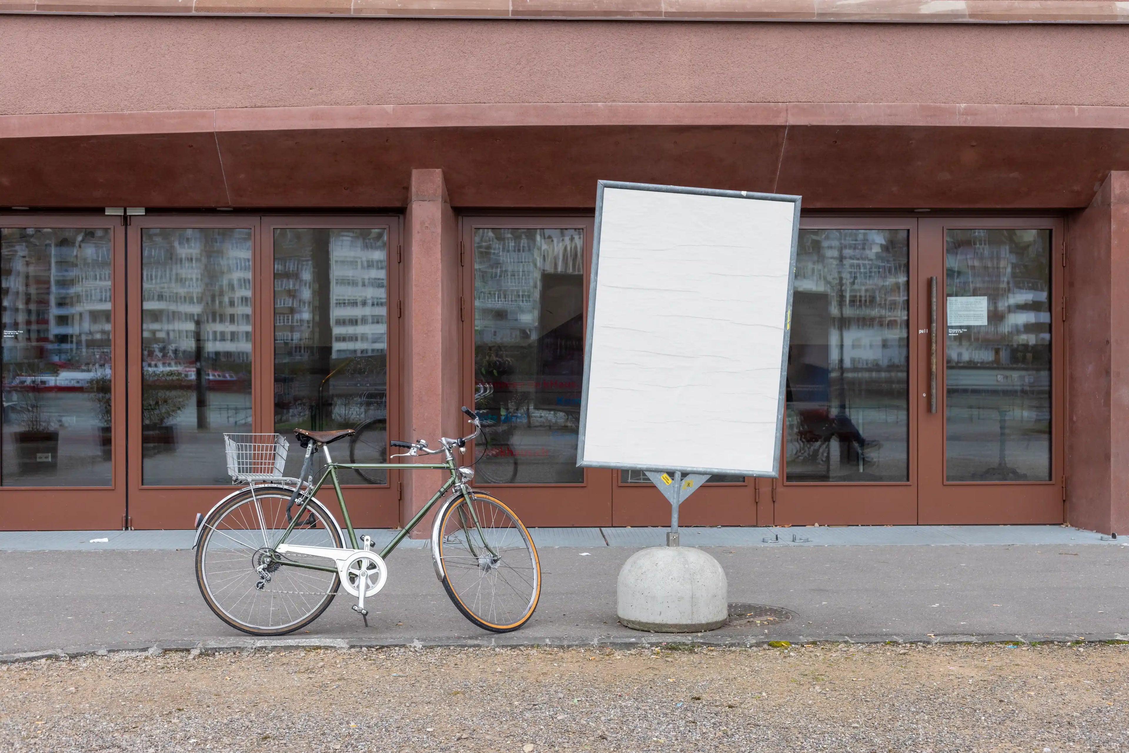 Blank white DIN poster mockup on a freestanding metal display at Kaserne Basel near the Rhine. A bicycle is parked beside the sign, with reflective glass doors, reddish-brown building details, gravel ground and a quiet urban riverside atmosphere.