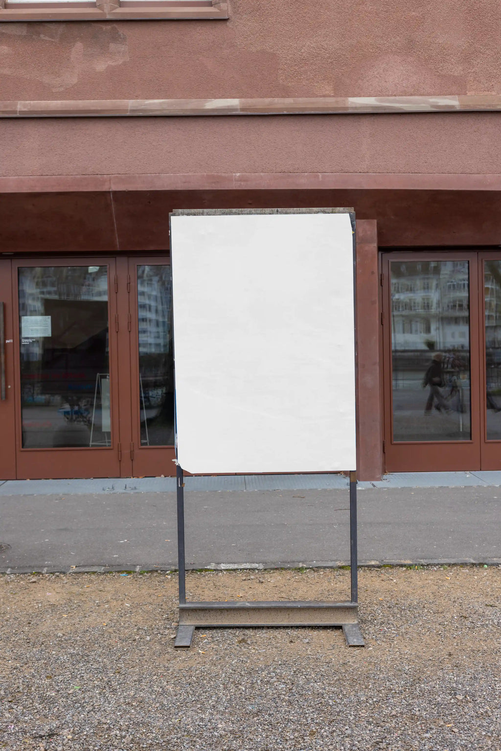 Blank white vertical poster mockup on a freestanding outdoor sign display at Kaserne Basel near the Rhine. The scene includes a metal sign frame, gravel ground, reddish-brown entrance doors, glass reflections and a realistic cultural venue environment.
