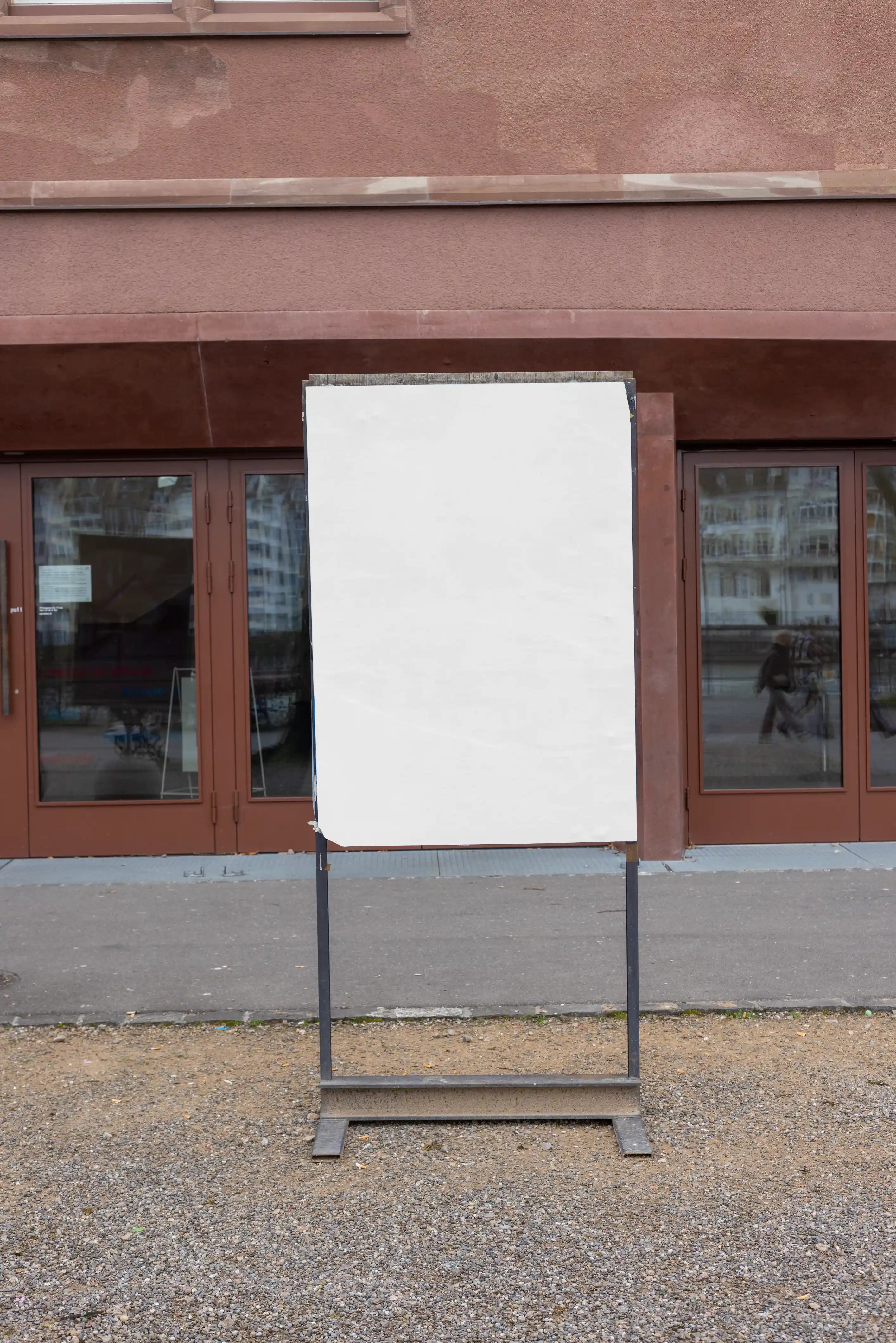 Blank white vertical poster mockup on a freestanding outdoor sign display at Kaserne Basel near the Rhine. The scene includes a metal sign frame, gravel ground, reddish-brown entrance doors, glass reflections and a realistic cultural venue environment.