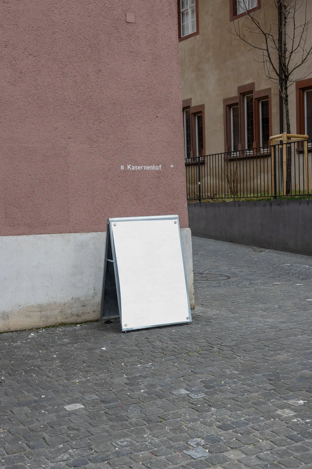 Blank white DIN poster mockup attached to a freestanding magnetic board with metal corner magnets at Kasernenhof in Basel. The scene shows cobblestone pavement, a pink textured wall, surrounding historic building details and a quiet courtyard atmosphere.