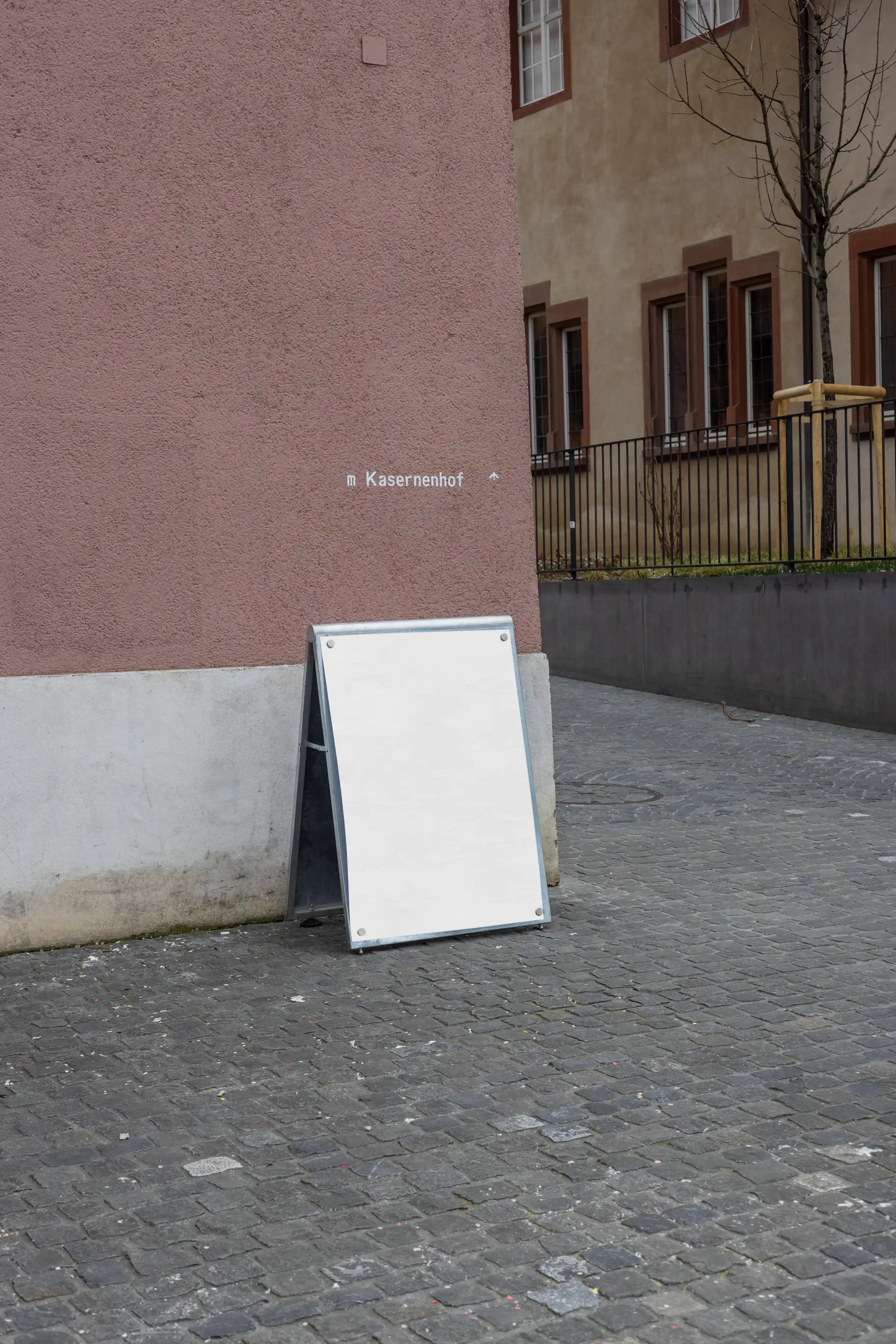 Blank white DIN poster mockup attached to a freestanding magnetic board with metal corner magnets at Kasernenhof in Basel. The scene shows cobblestone pavement, a pink textured wall, surrounding historic building details and a quiet courtyard atmosphere.