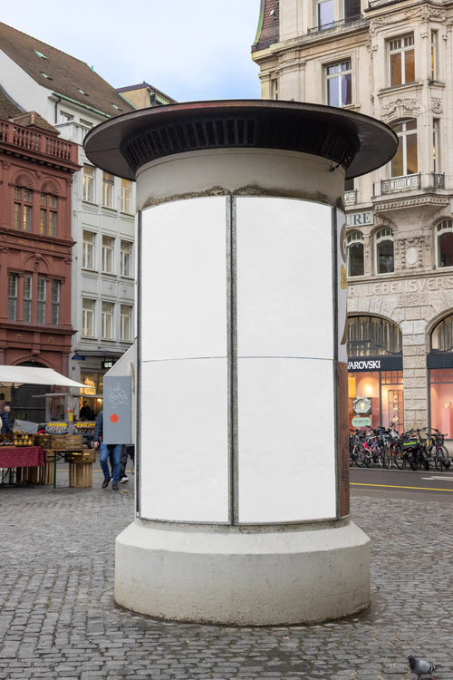 Blank white cylindrical ad column mockup at Basel Marktplatz with four vertical poster areas on a curved surface. The location includes historic architecture, cobblestone pavement, bicycles and street details, created for realistic Litfaßsäule, poster campaign and out-of-home advertising previews.