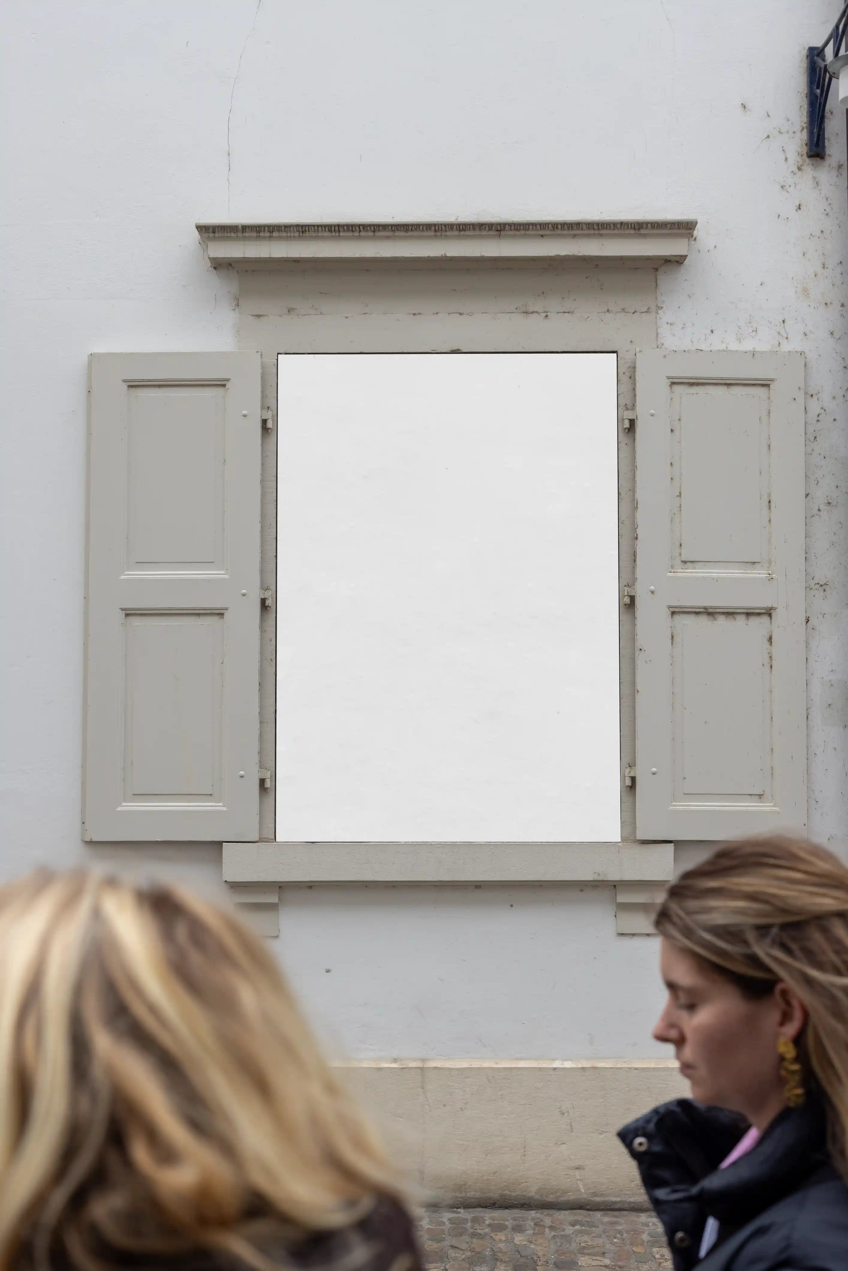 Blank white DIN poster mockup in a window display at the Museum of Cultures Basel near Münsterplatz. The image includes pale shutters, a white facade, subtle wall texture and two blurred people walking in the foreground for a natural city atmosphere.