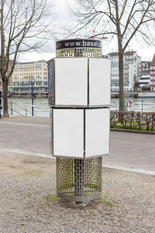 Four blank white DIN poster mockups on a cylindrical advertising column near the Rhine riverside in Basel. The scene shows a modern metal ad column on gravel, bare trees, city buildings, water and natural outdoor light for realistic out-of-home poster presentations.
