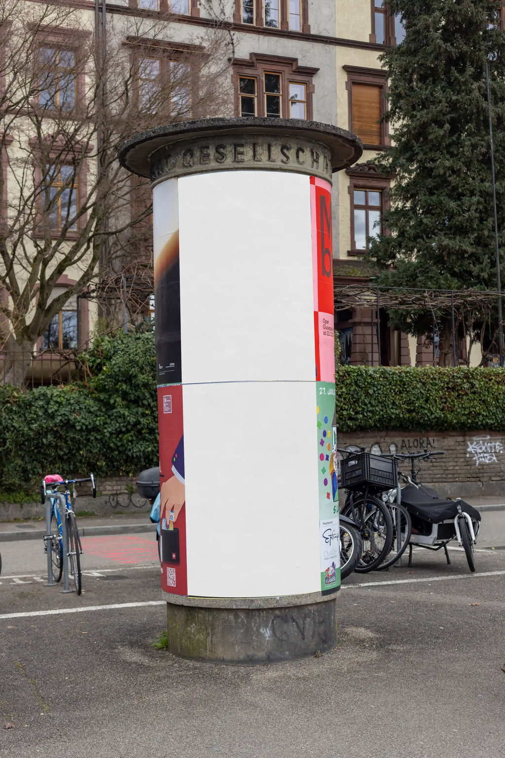Blank white two-part poster mockup on a cylindrical advertising column near the Rhine in Basel. The location includes bicycles, trees, city buildings and street textures, perfect for DIN poster designs, Litfaßsäule mockups and realistic outdoor advertising previews.
