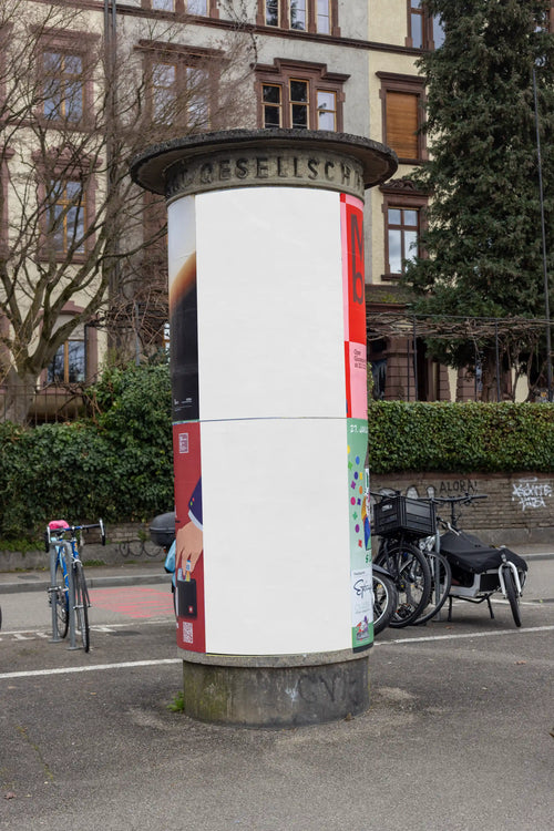 Blank white two-part poster mockup on a cylindrical advertising column near the Rhine in Basel. The location includes bicycles, trees, city buildings and street textures, perfect for DIN poster designs, Litfaßsäule mockups and realistic outdoor advertising previews.
