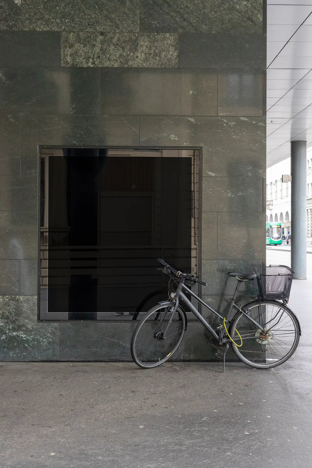 Blank dark square city light display mockup in Basel set into a polished stone wall, with a bicycle parked in front of the glass frame. The urban scene includes reflections, pavement, a covered walkway and a green tram in the distance.