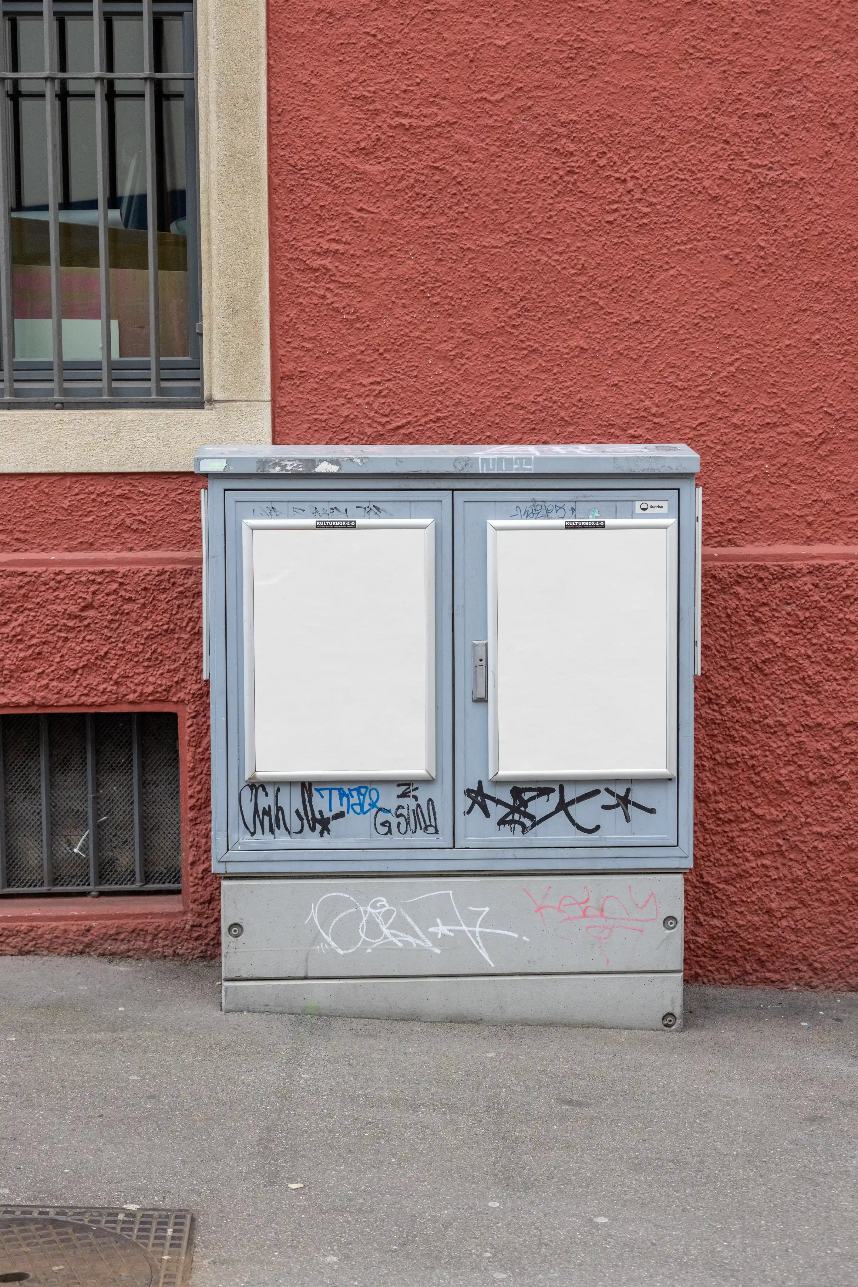 Two blank white DIN poster mockups in silver frames on a grey utility box in Basel. The scene shows graffiti, weathered metal, a textured red wall and a barred window, giving the mockup an authentic city street look.