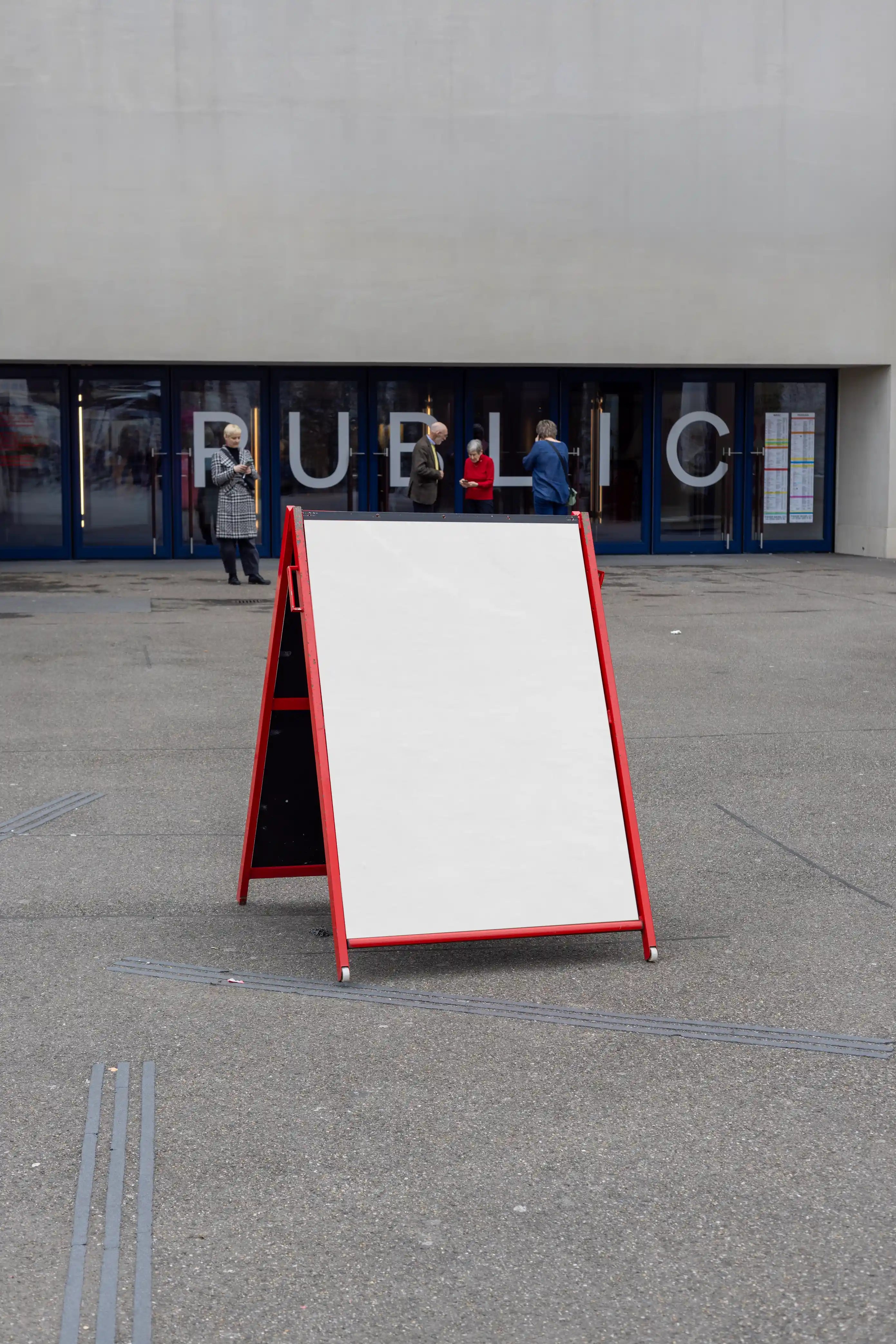 Blank white A-frame poster mockup with a red frame outside a public entrance near Theater Basel. The display stands on grey pavement, with large glass doors, people in the background and a clean cultural venue setting behind it.