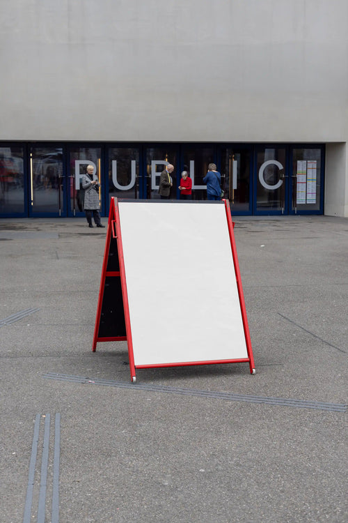 Blank white A-frame poster mockup with a red frame outside a public entrance near Theater Basel. The display stands on grey pavement, with large glass doors, people in the background and a clean cultural venue setting behind it.