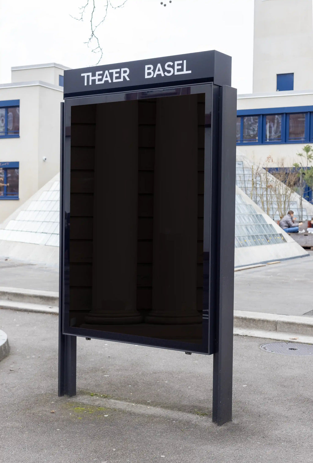 Blank dark DIN format city light poster mockup in front of Theater Basel, set inside a black outdoor display frame. The image includes the Theater Basel sign, modern building details, glass pyramid structures, pavement and an authentic cultural city setting.