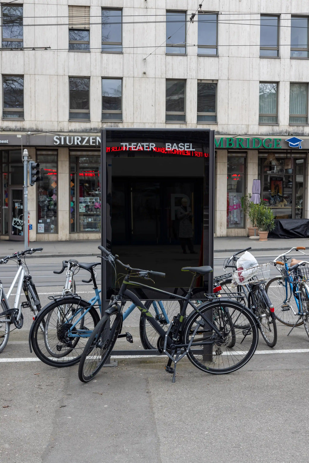 Blank dark DIN poster mockup inside a black city light display near Theater Basel. Multiple bicycles stand in front of the display, with street shops, tram wires, reflections and an authentic Basel out-of-home advertising environment around it.