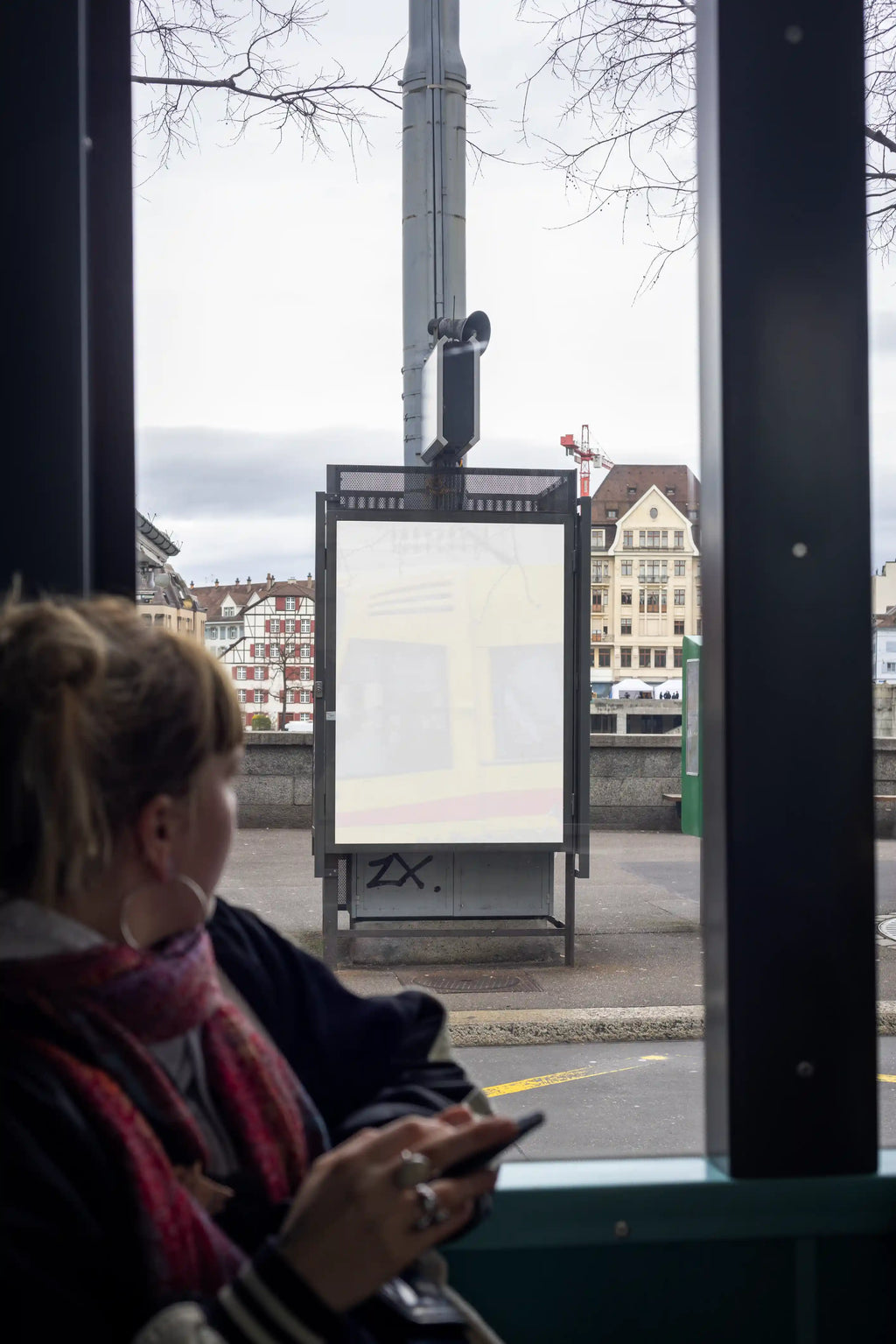 Blank pale DIN poster mockup viewed from inside a tram in Basel near Mittlere Brücke. A blurred passenger sits in the foreground, while the poster display, street, historic buildings and riverside area are visible through the tram window.