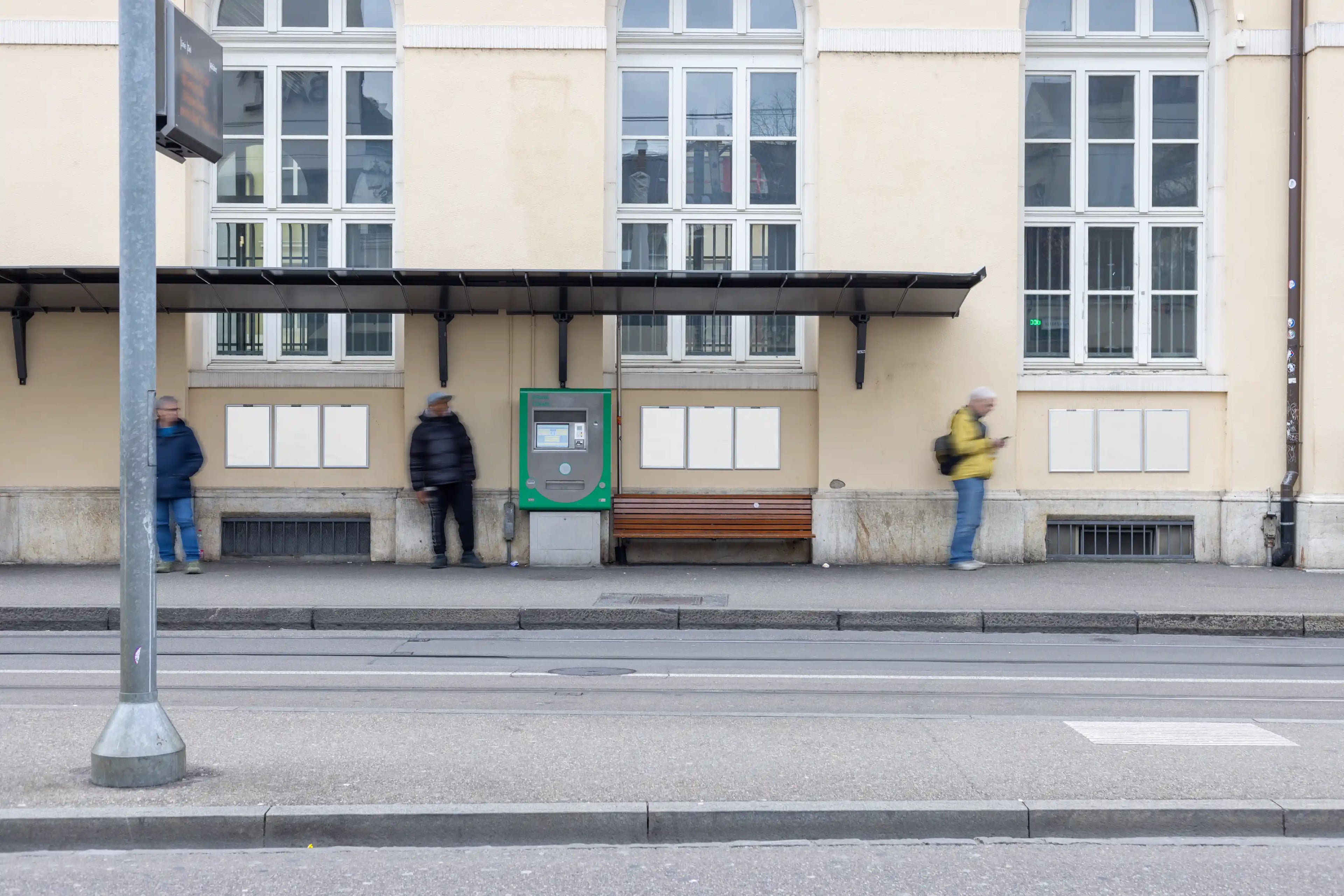 Nine blank white DIN poster mockups arranged in three groups on a tram stop wall in Basel. A bench, ticket machine, blurred people, street tracks and tall windows create a realistic public transport setting for out-of-home poster presentations.