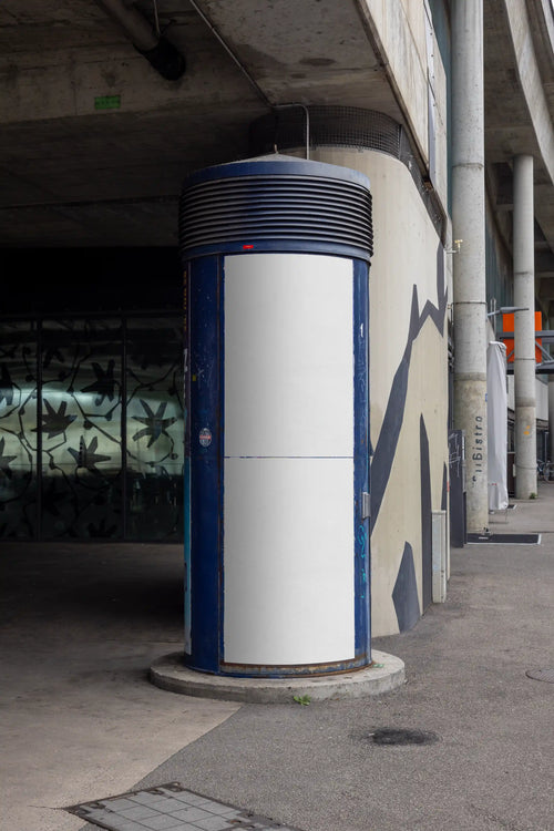 Blank white two-part poster mockup on a curved blue advertising column in Basel under a concrete bridge structure. The urban setting includes worn surfaces, shadows, concrete architecture and street surroundings, ideal for Litfaßsäule mockups, DIN poster previews and outdoor advertising concepts.