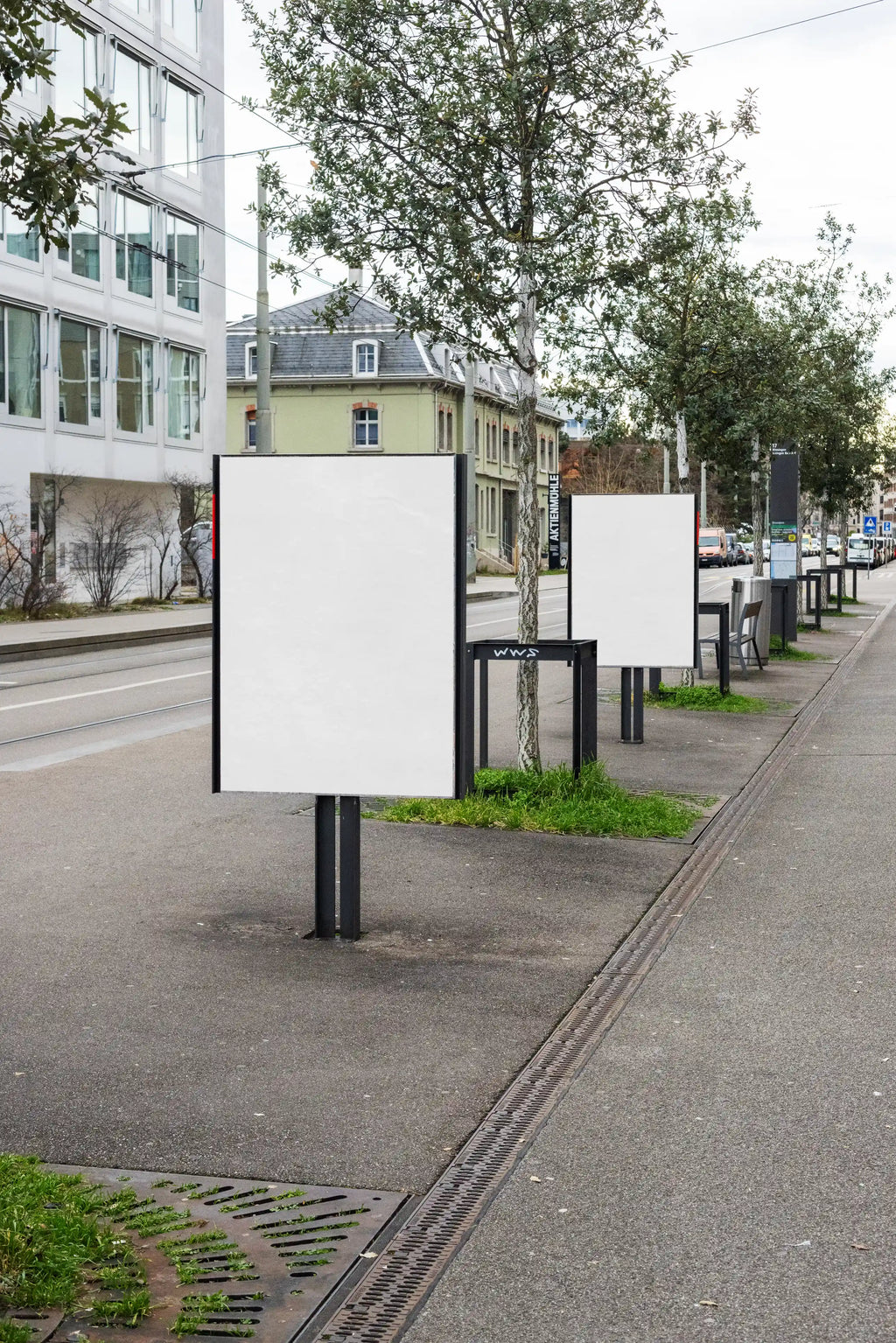 Two blank white DIN poster mockups on freestanding outdoor poster boards at Wiesenplatz in Basel. The scene shows a city sidewalk with trees, tram tracks, grass, street details and buildings, creating a realistic urban out-of-home advertising setting.
