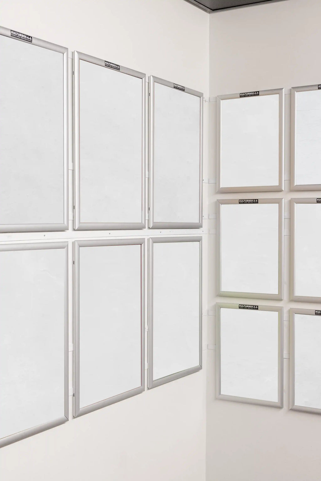 Twelve blank white DIN poster mockups displayed in silver frames across two white indoor walls. The frames are arranged in a clean corner gallery layout, making the scene suitable for presenting a full poster series, campaign or art collection.