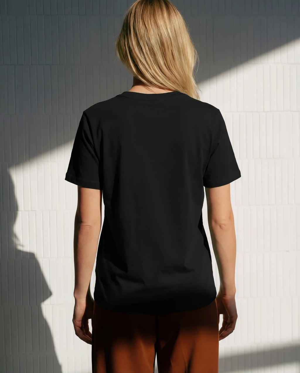 Female black t-shirt mockup from the back with blank editable shirt surface, natural daylight, realistic fabric texture and clean wall background, perfect for clothing brands, apparel mockups, back designs and streetwear presentations.