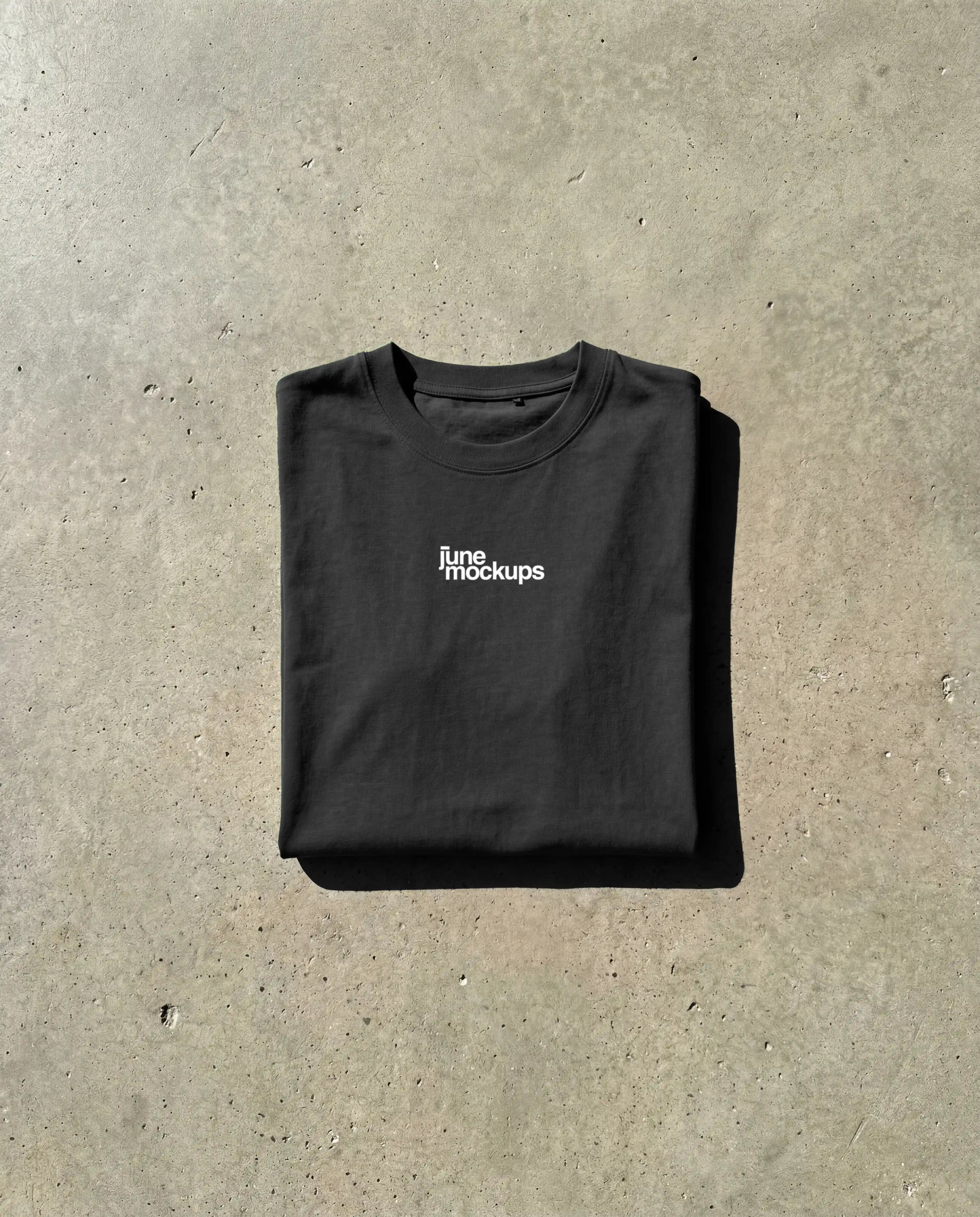 Free black t-shirt mockup folded flat lay with small chest logo, realistic cotton fabric texture, natural daylight shadows on raw concrete background, high resolution apparel mockup free download for streetwear branding, fashion design and e-commerce product presentation
