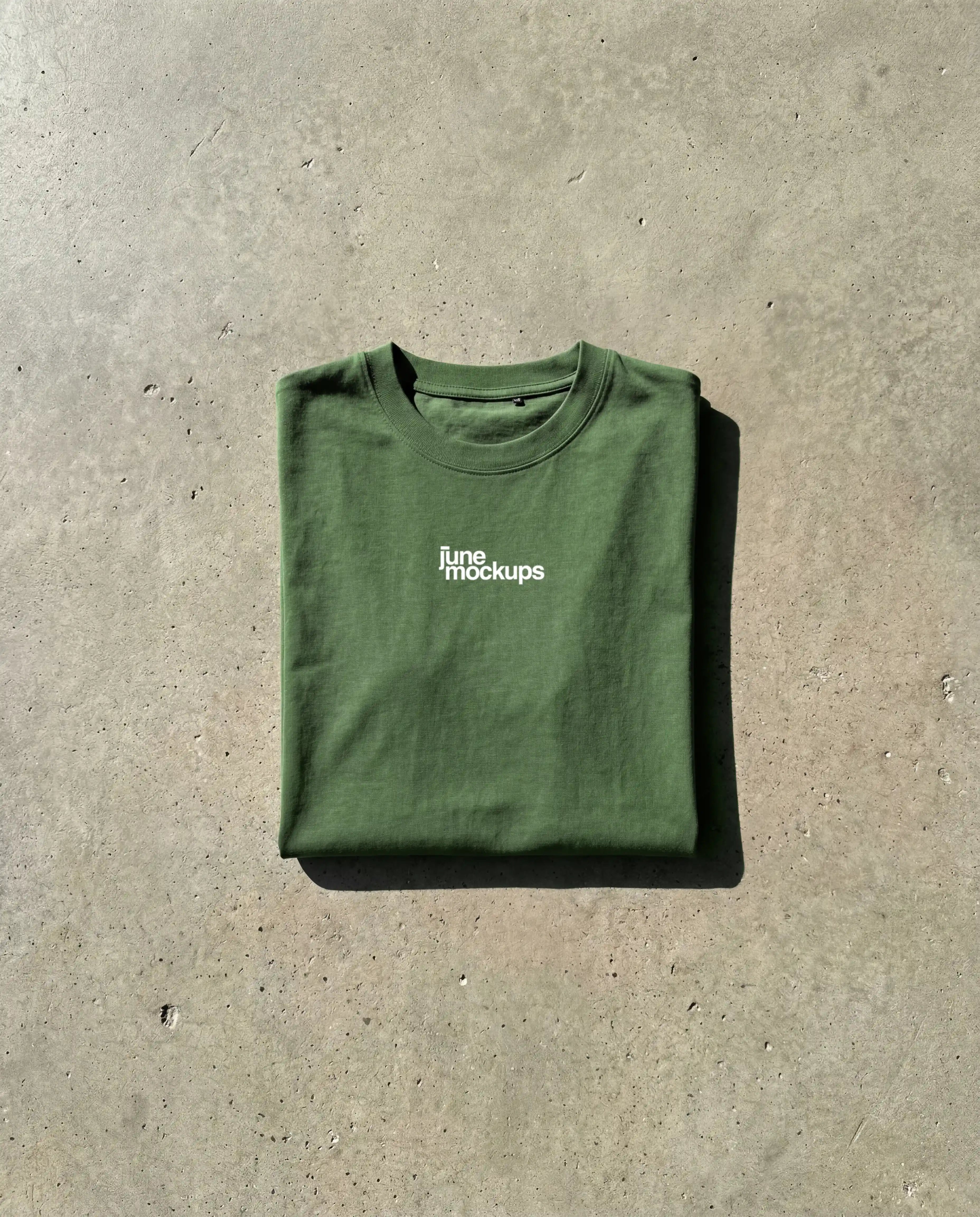 Free green t-shirt mockup folded flat lay with centered logo print, realistic fabric folds and natural lighting on textured concrete surface, high quality apparel mockup free download for designers, clothing brands and online shop visuals