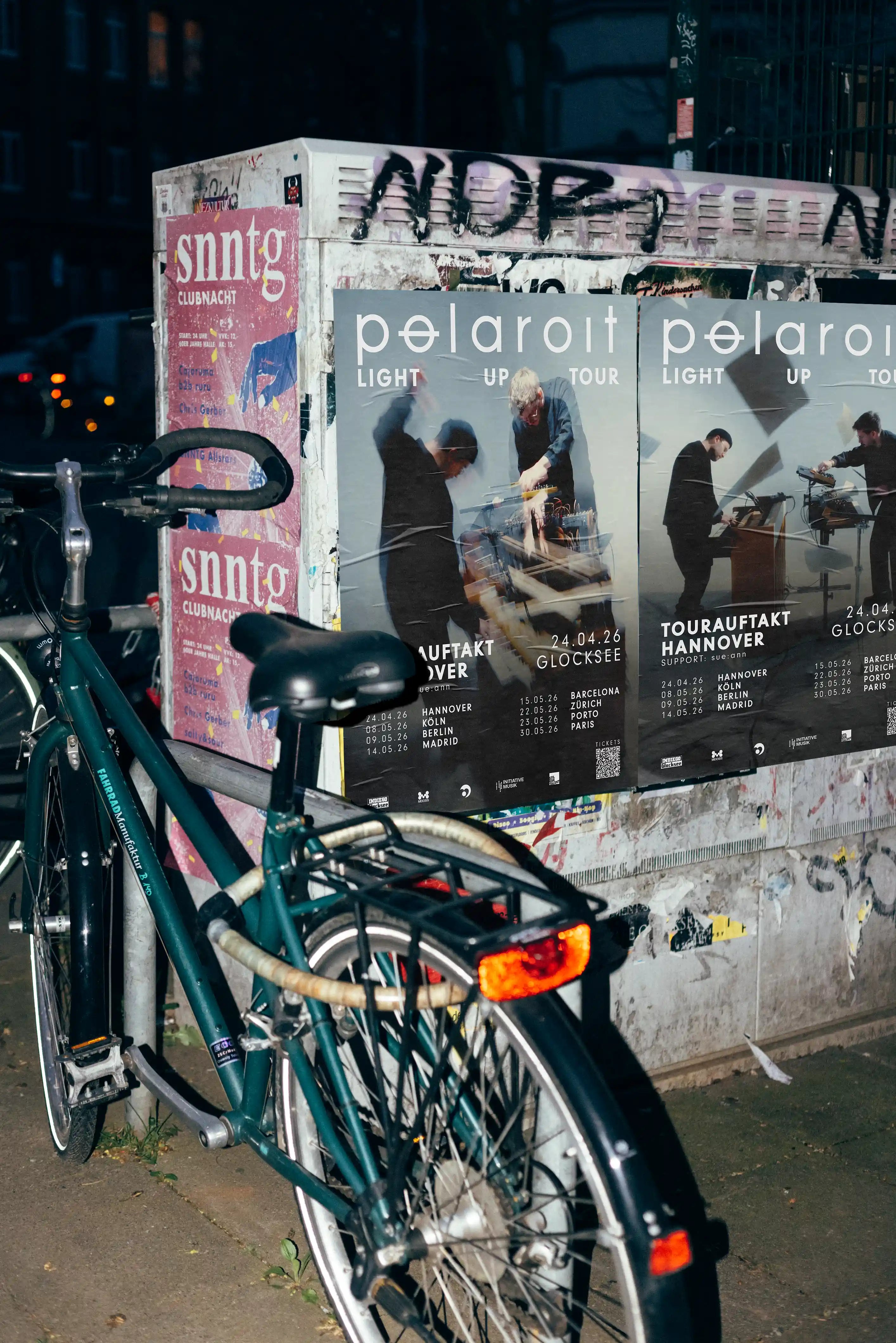 Night flash urban poster mockup showing realistic street advertising posters of Polaroit Light Up Tour placed on a textured wall next to a bicycle in an urban environment, captured with direct flash for strong shadows and authentic city atmosphere.